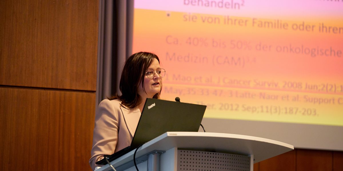 Frau am Rednerpult, hält Vortrag mit Projektion im Hintergrund; Text erwähnt Onkologie und Komplementärmedizin.