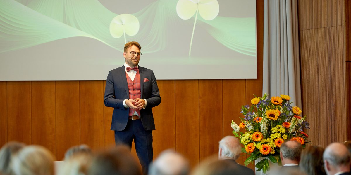 Ein Mann im Anzug hält einen Vortrag auf einer Bühne vor Publikum, daneben steht ein bunter Blumenstrauß.