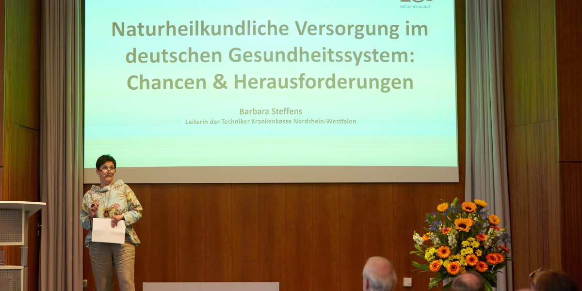 Eine Frau spricht vor einem Publikum über naturheilkundliche Versorgung im deutschen Gesundheitssystem.