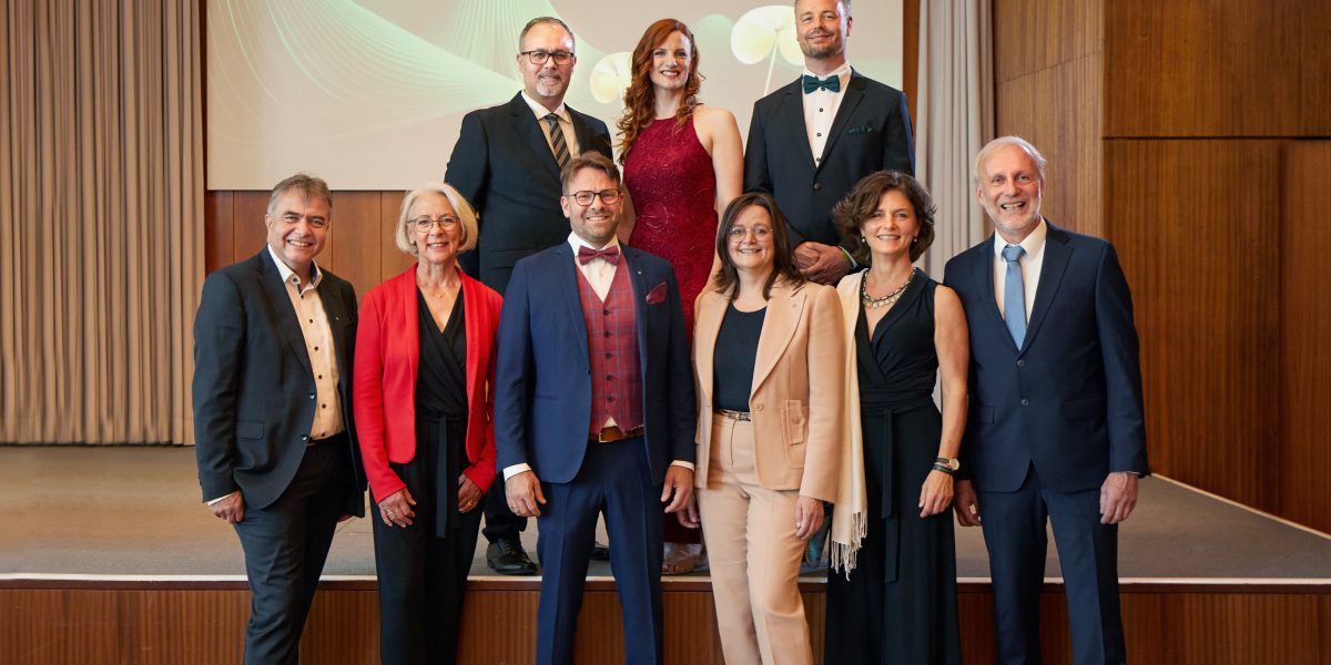 Neun elegant gekleidete Personen stehen in einem Saal vor einem Banner mit der Aufschrift