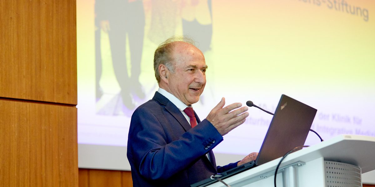 Ein Mann hält eine Rede an einem Pult vor einer Leinwand in einem Holzvertäfelten Raum.