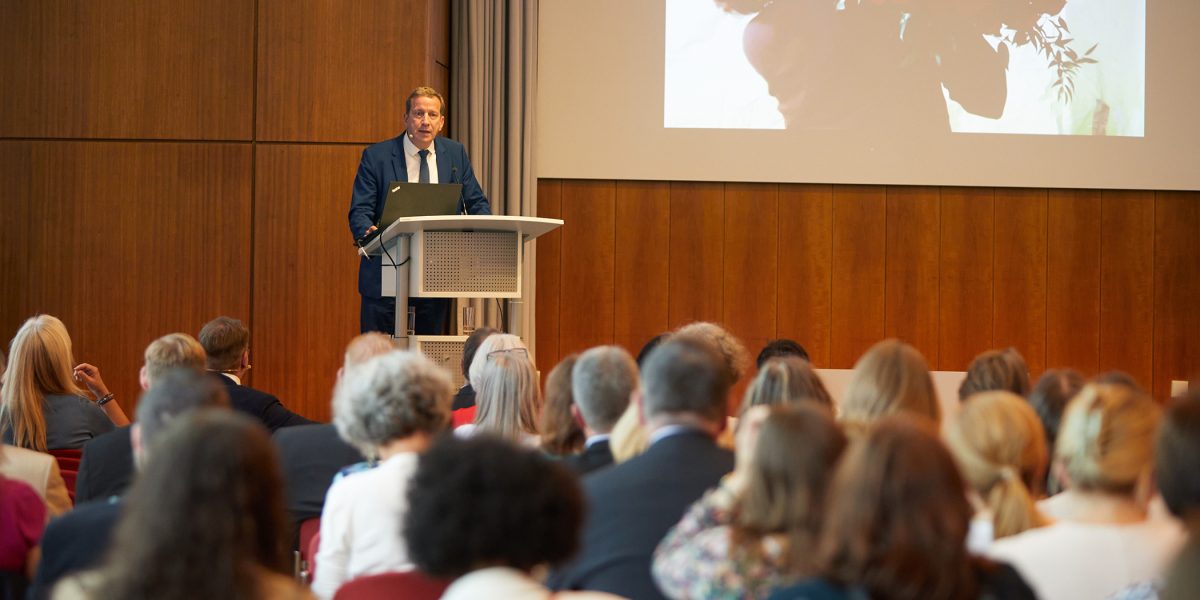 Ein Mann hält einen Vortrag vor Publikum, auf der Leinwand ein Bild mit einer Person und einem großen Rosengesteck.