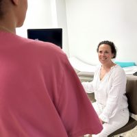 Frau in weißer Kleidung sitzt lächelnd in Arztpraxis; Ärztin steht im Vordergrund mit Monitor im Hintergrund.