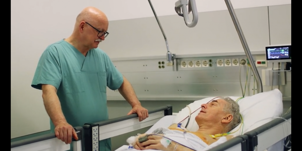 Ein Arzt spricht mit einem älteren Patienten, der in einem Krankenhausbett liegt; medizinische Geräte im Hintergrund.