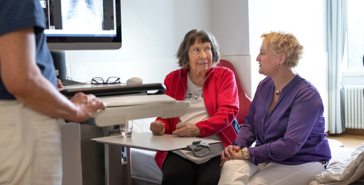 Zwei Frauen sitzen in einem Krankenhauszimmer und sprechen mit einem Arzt. Auf einem Monitor sind Röntgenbilder zu sehen.
