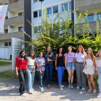 Neun Frauen stehen gemeinsam lächelnd vor einem Gebäude neben einem Schild mit der Aufschrift 