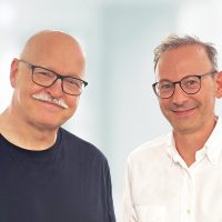 Zwei Männer mit Brillen lächeln in die Kamera, der eine trägt ein schwarzes T-Shirt, der andere ein weißes Hemd.