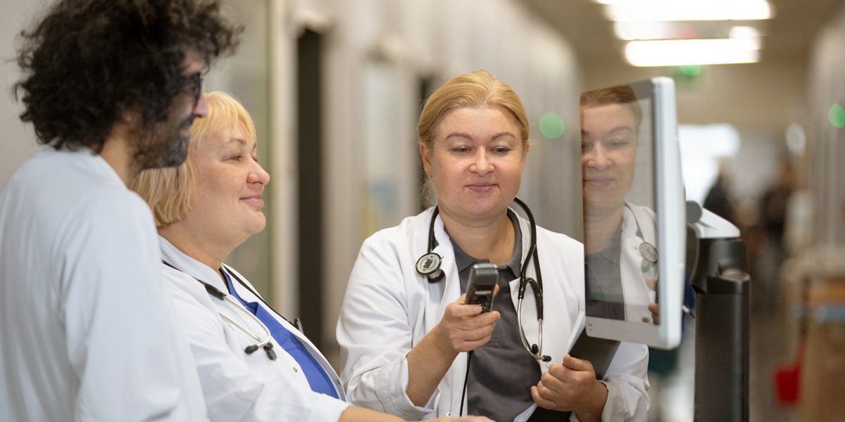Drei Ärzt:innen in weißen Kitteln betrachten gemeinsam einen Monitor in einem Krankenhausflur.