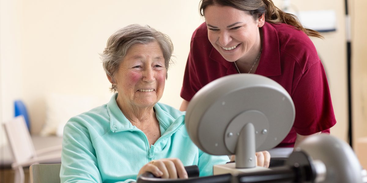 Eine ältere Frau trainiert auf einem Ergometer, während eine Pflegerin sie lächelnd unterstützt.