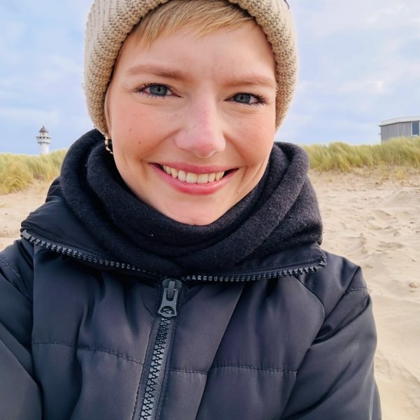 Frau mit Mütze und Jacke lächelt am Strand, im Hintergrund Gräser und ein Leuchtturm.