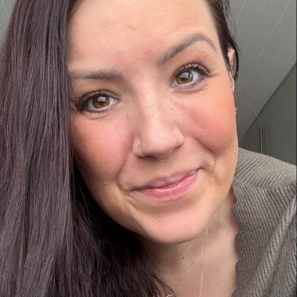 Frau mit dunkelbraunen Haaren und Nasenpiercing lächelt freundlich in die Kamera. Sie trägt einen grauen Pullover.