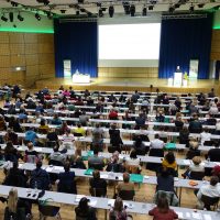 Voller Konferenzsaal mit Publikum, das einem Vortrag auf einer Bühne mit Projektor folgt.