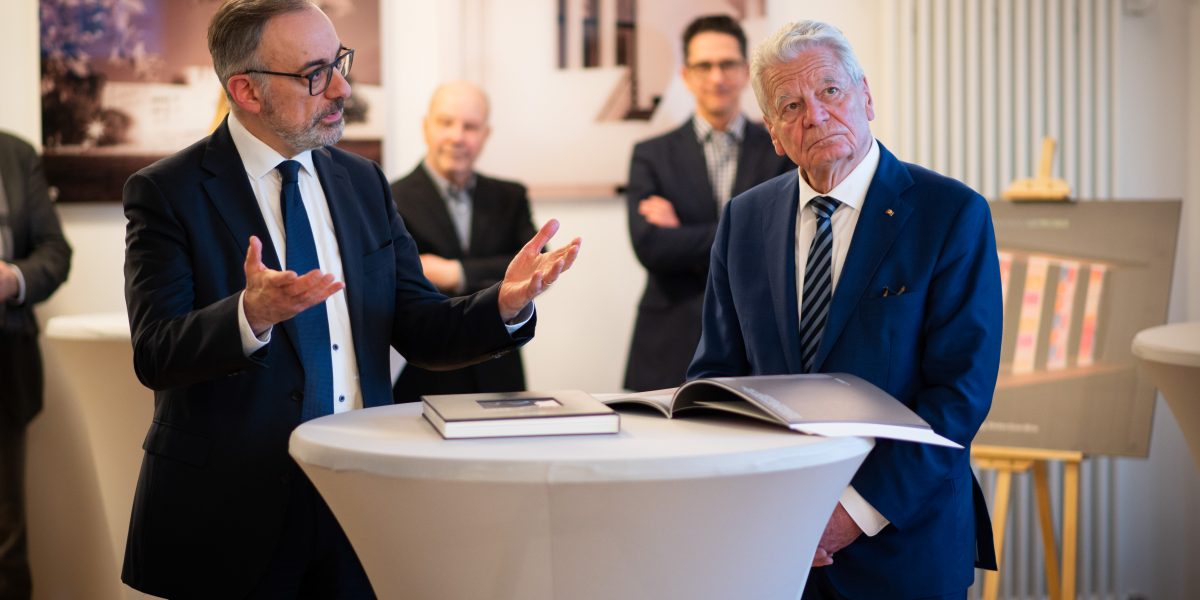 Zwei Männer in Anzügen stehen an einem runden Tisch, einer spricht gestikulierend, der andere hört zu, ein Buch liegt offen.