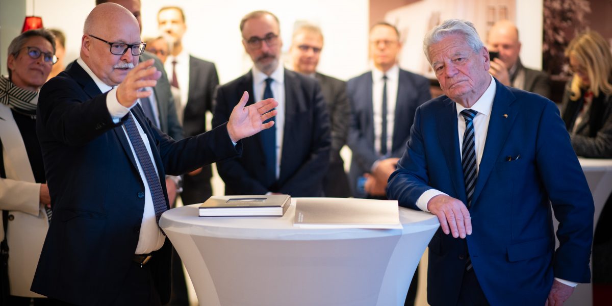 Älterer Mann gestikuliert beim Reden, während ein anderer Mann neben ihm an einem Stehtisch steht, umgeben von Zuhörern.