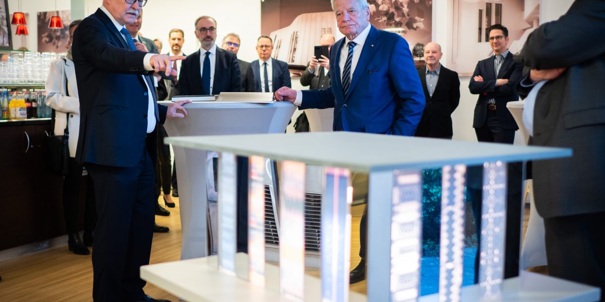 Gruppe von Männern in Anzug um einen Tisch, einer zeigt auf ein architektonisches Modell im Vordergrund.