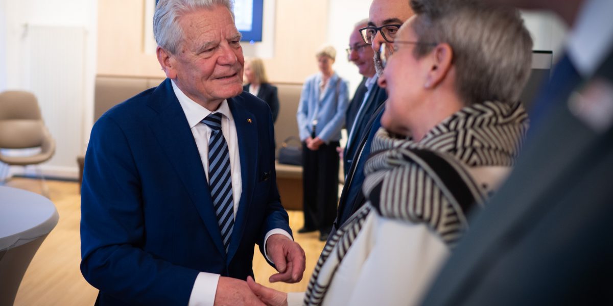 Älterer Mann im blauen Anzug schüttelt einer Frau die Hand. Mehrere Personen im Hintergrund in einem Raum.