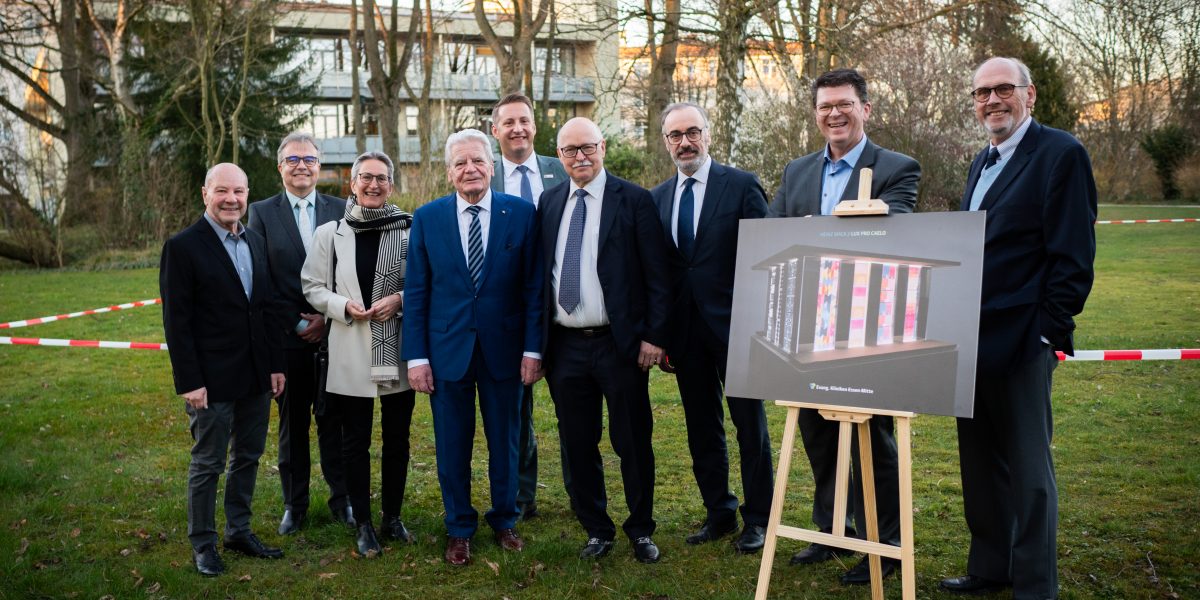 Eine Gruppe von Menschen in formeller Kleidung steht in einem Park neben einem Plakat auf einer Staffelei.