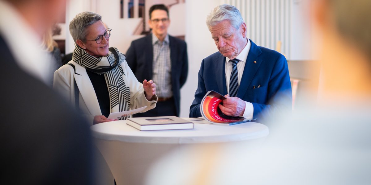 Zwei ältere Personen stehen an einem Tisch mit Büchern. Eine spricht lächelnd, die andere blättert interessiert in einem Buch.