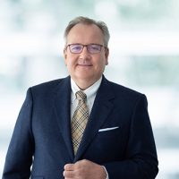 Älterer Mann mit Brille im Anzug, lächelnd vor unscharfem, hellem Hintergrund.