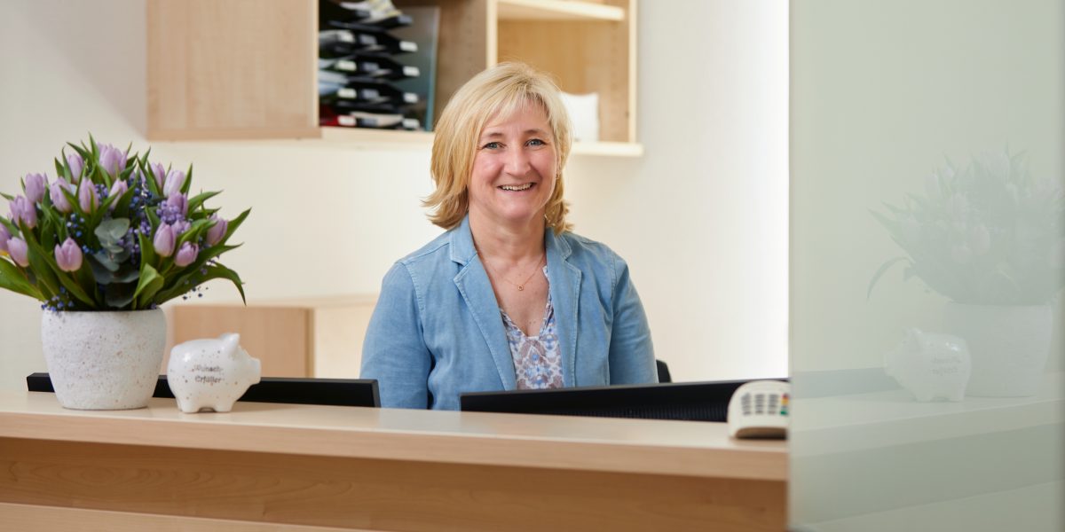 Frau in blauem Blazer lächelt an einem Empfangstresen mit Blumenstrauß und Dekoration.