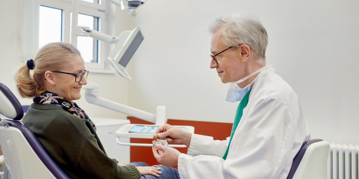 Zahnarzt zeigt einer lächelnden Patientin in einem Behandlungsraum ein Zahnmodell.