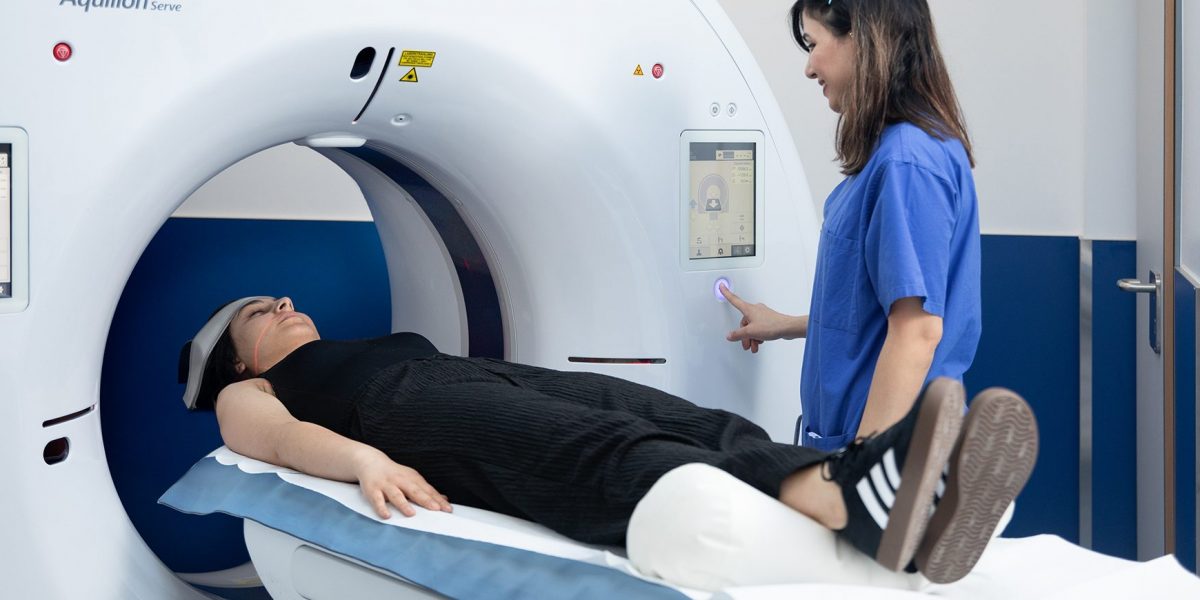 Patientin liegt in einem CT-Scanner, während eine medizinische Fachkraft in blauer Kleidung den Scanner bedient.