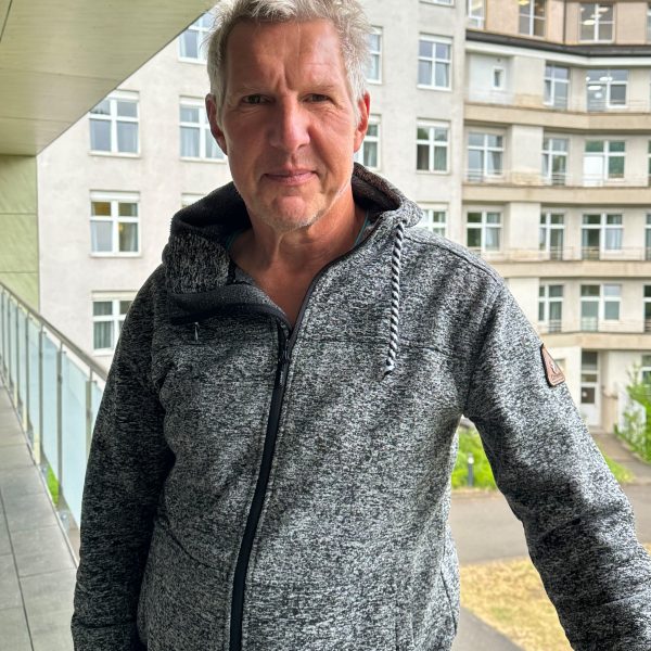 Mann in grauem Kapuzenpullover steht auf einem Balkon vor einem Gebäude mit vielen Fenstern.