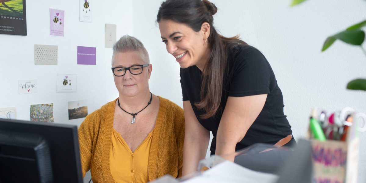 Zwei Frauen lächeln am Computer, die Wand ist mit Karten dekoriert. Eine sitzt, die andere steht neben ihr.