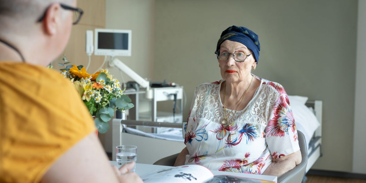 Frau mit Kopftuch und Brille sitzt in einem Krankenhauszimmer, gegenüber einer anderen Person, ein Magazin liegt auf dem Tisch.