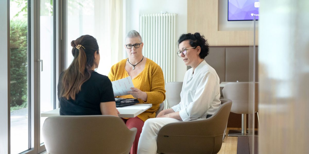 Drei Personen sitzen an einem Tisch in einem Gespräch. Eine Person hält ein Dokument, die anderen hören aufmerksam zu.