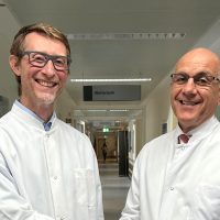 Zwei Männer im weißen Kittel schütteln sich lächelnd die Hand in einem Krankenhausflur.