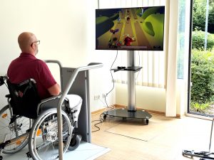 Ein Mann im Rollstuhl spielt ein Videospiel auf einem großen Bildschirm in einem hellen Raum.