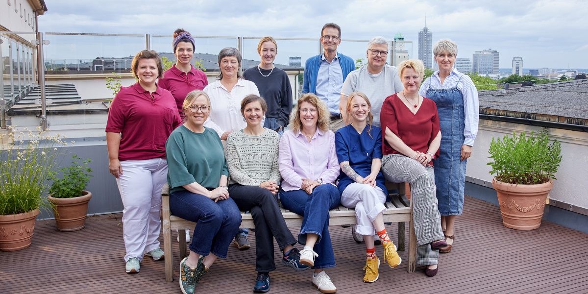 Eine Gruppe von 13 Personen posiert auf einer Dachterrasse mit Pflanzen und Stadtblick im Hintergrund.
