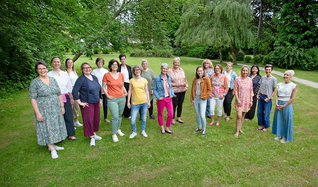 Gruppe von Frauen steht auf einer Wiese im Park, umgeben von Bäumen. Sie lächeln in die Kamera.