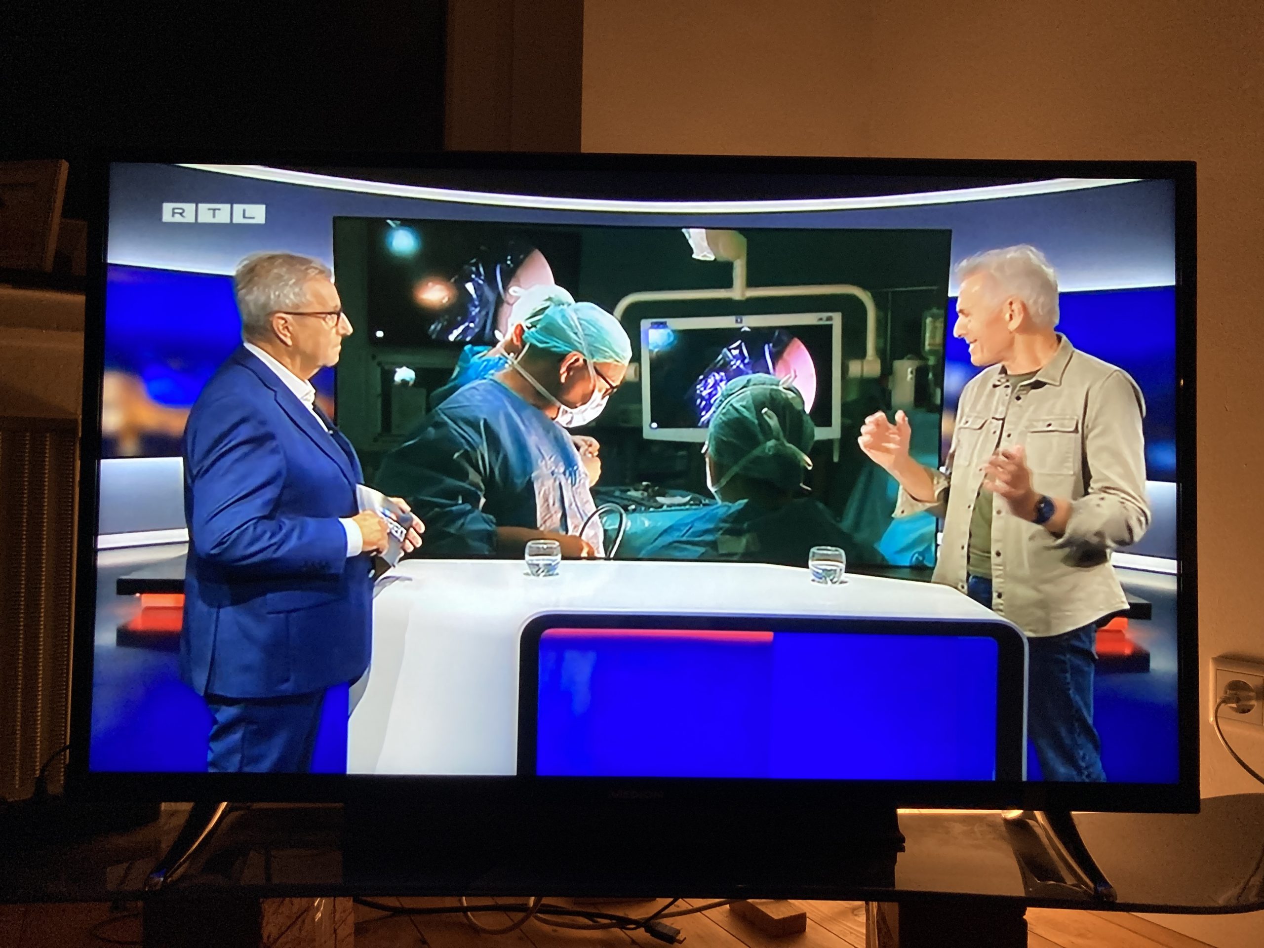 Zwei Männer auf einem RTL-Set diskutieren vor einem Bild eines OP-Teams auf einem Bildschirm.