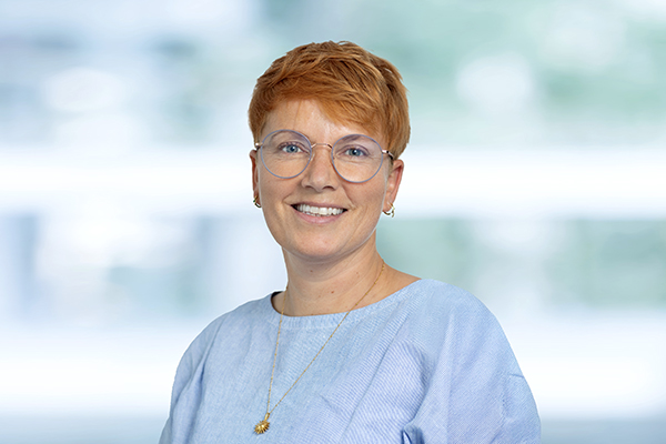 Frau mit kurzem, rotbraunem Haar, Brille und blauem Oberteil lächelt vor verschwommenem Hintergrund in die Kamera.