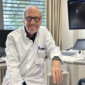 Ein älterer Mann in weißem Arztkittel lächelt in einer Praxis, neben einem Ultraschallgerät.