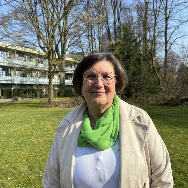 Ältere Frau mit Brille und grünem Schal steht im Park vor einem Gebäude, Bäume im Hintergrund, blauer Himmel.