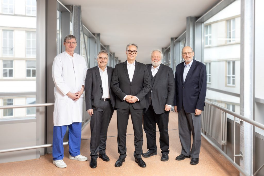 Fünf Männer, einer in Arztkleidung, stehen in einem modernen, lichtdurchfluteten Korridor und lächeln in die Kamera.