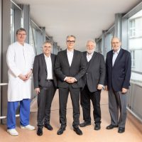 Fünf Männer, einer in Arztkleidung, stehen in einem modernen, lichtdurchfluteten Korridor und lächeln in die Kamera.