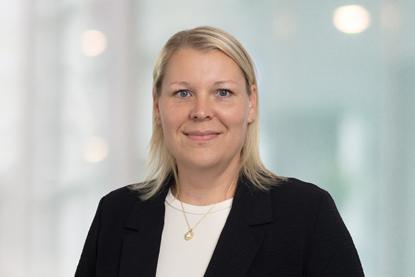 Porträt einer lächelnden Frau mit blondem Haar in einem schwarzen Blazer vor einem unscharfen, hellen Hintergrund.