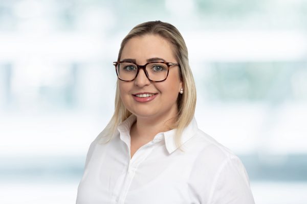 Junge Frau mit Brille und blonder Frisur in weißer Bluse lächelt vor unscharfem Hintergrund in die Kamera.