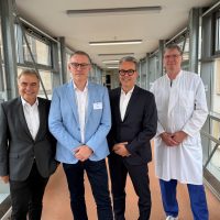 Vier Männer in Anzügen und Arztkittel stehen in einem Korridor mit Fenstern und Metallstruktur.